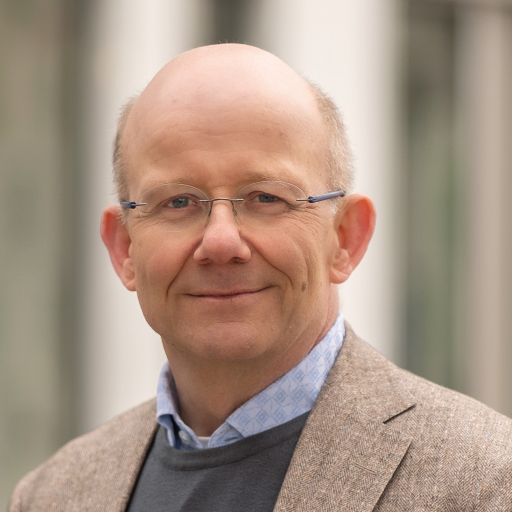 Porträtfoto von Dr. Stefan Breuer. Zu sehen ist ein Mann mittleren Alters mit einer Glatze und einer schmalen, randlosen Brille. Er trägt ein hellbraunes Tweed-Sakko über einem dunkelgrauen Pullover und einem hellblauen Hemd mit dezenten Mustern. Er lächelt ruhig und freundlich in die Kamera.